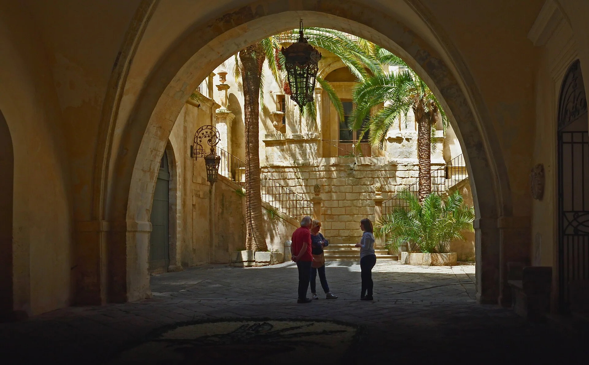 Cortile di un palazzo a Noto, con archi, palme e persone